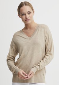 Beige, könnyű V nyakú pulóver hosszú ujjal, bordázott mandzsettával és szegéllyel, finom textúrával és logóval az oldalon.