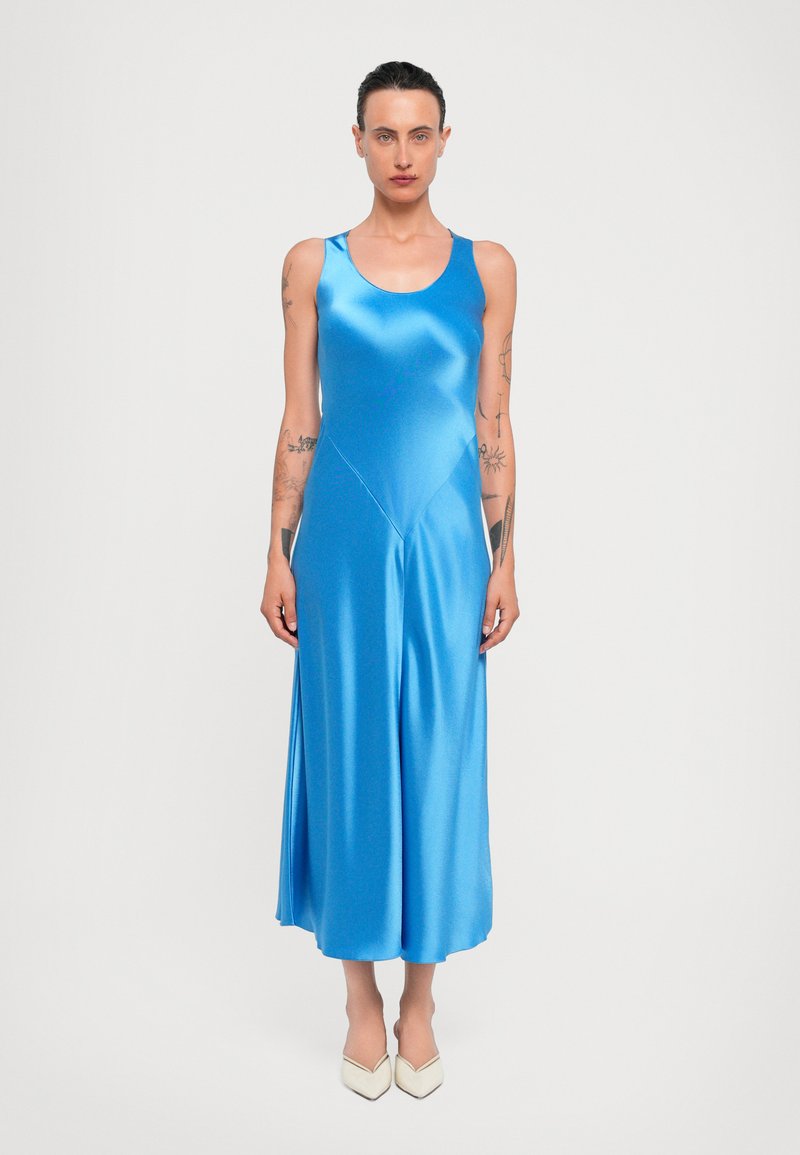 Satin blauwe midi-jurk met een ronde hals, aansluitende top en uitlopende rok. Gladde textuur en minimalistisch ontwerp. Gemaakte witte hakken.