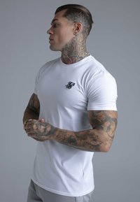 Hvid tætsiddende T-shirt med korte ærmer, rund hals og lille sort broderet logo på brystet. Glat bomuldsmateriale, minimalistisk design.