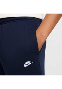 A sötétkék Nike melegítőnadrág textúrázott anyagból készült, oldalsó zsebekkel rendelkezik, és a bal combon fehér Nike logó van hímezve.