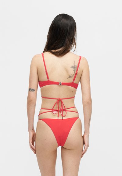 Γυναίκα με μακριά σκούρα μαλλιά που φοράει κόκκινο μπικίνι με πολλαπλές λωρίδες δεμένες πίσω, εμφανίζοντας τατουάζ στην άνω πλάτη και το αριστερό χέρι.