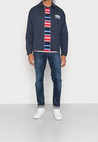 Marinblå bomberjacka med dragkedja framtill, vitt märke, randig röd och blå t-shirt under, blå jeans och vita sneakers.