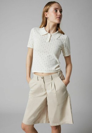 Nuori nainen, jolla on kädet taskuissa, pukeutuneena valkoiseen lyhythihaiseen neulepoliopaitaan ja beigeihin polvipituisiin laskoshousuihin.