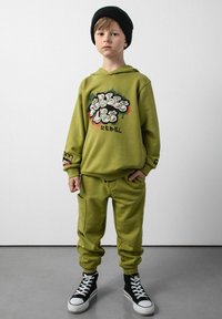 Oliwkowa bluza z kapturem z grafiką w stylu graffiti, w połączeniu z pasującymi dresami z kieszeniami. Czarna czapka beanie i sneakersy za kostkę dopełniają wygląd.