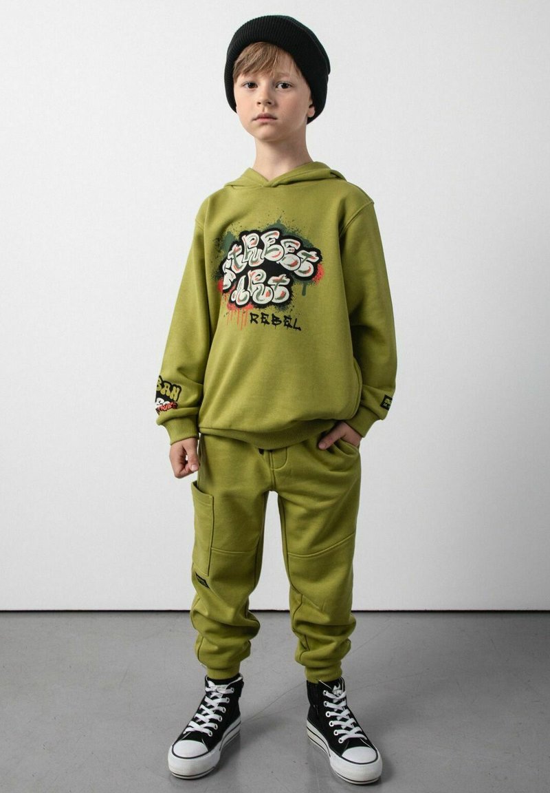 Oliwkowa bluza z kapturem z grafiką w stylu graffiti, w połączeniu z pasującymi dresami z kieszeniami. Czarna czapka beanie i sneakersy za kostkę dopełniają wygląd.