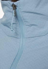 Veste zippée bleu clair avec col montant, présentant une surface texturée et un zip central avec un tirette lisse et un cordon de serrage.