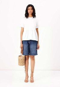 Hvit kortermet peplumtopp kombinert med denim bermudashorts. En flettet tan håndveske og stroppede beige sandaler fullfører antrekket.
