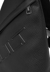 Zaino in pelle nera con superficie testurizzata e dettaglio logo in rilievo, chiusura con zip e forma triangolare strutturata.