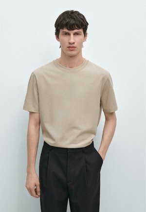 Giovane uomo con capelli corti e scuri che indossa una camicia beige a maniche corte infilata in pantaloni neri a vita alta, in piedi con una mano in tasca.