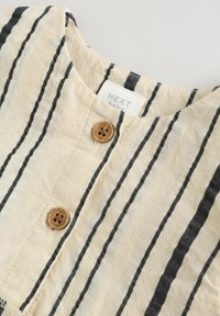 Vêtement pour bébé rayé en beige clair et noir. Présente un col rond, deux boutons en bois et une étiquette indiquant "NEXT baby".