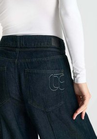 Donkerblauwe denim jeans met een hoge tailleband. Beschikt over twee achterzakken, waarvan er één witte geborduurde details heeft, met een gestructureerde stof.