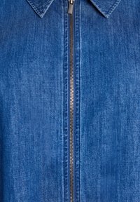 Närbild av en blå denimjacka med krage och metallblixtlås, vilket framhäver tygets struktur och färgvariationer.