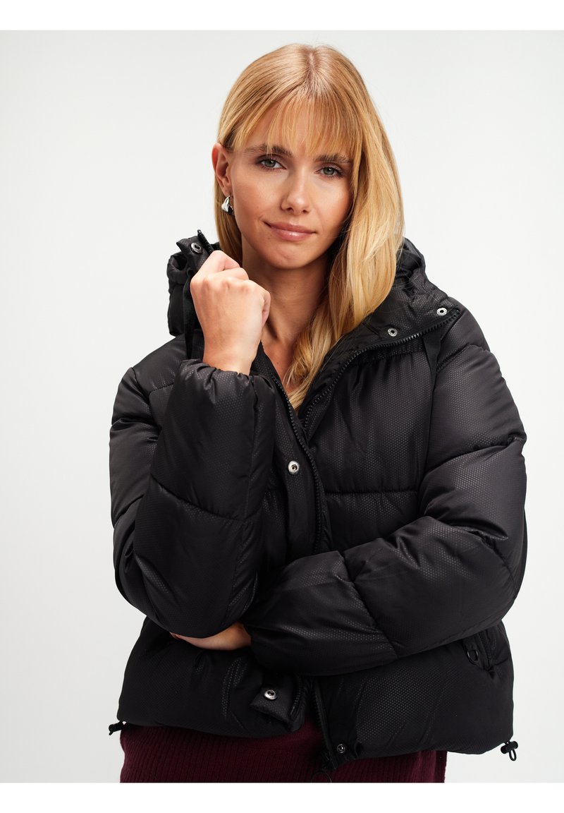 Blondynka w czarnej kurtce puchowej z kapturem, trzymająca kołnierz kurtki jedną ręką, na tle jasnego, jednolitego tła.