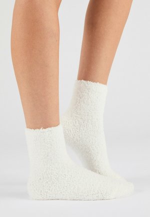 Paire de jambes portant des chaussettes cheville blanches et duveteuses sur un fond clair et uni.