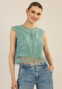 Ein mintgrünes Spitzen-Crop-Top mit gewellten Säumen, komplizierten Mustern und einem zarten Spitzenbesatz am Saum. Getragen mit blauen Jeans.