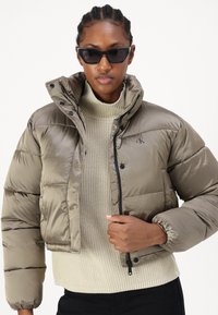 Giacca imbottita khaki con colletto alto, chiusura con zip e dettagli neri, indossata sopra un leggero maglione di lana. Presenta un logo sul petto.