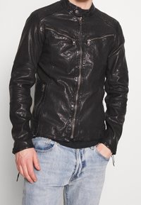 Giacca di pelle nera con colletto alto, tasche con zip e dettagli strutturati sulle spalle; presenta una vestibilità aderente e una finitura liscia e lucida.