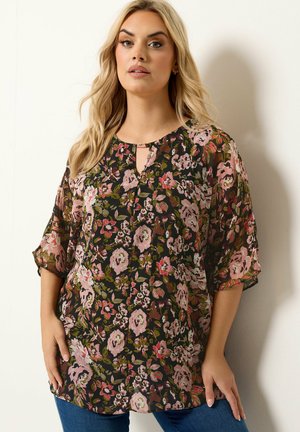 Lyshåret kvinde iført en sort blomstret bluse med pink og grønne blomster og kvartlange gennemsigtige ærmer, kombineret med blå jeans.