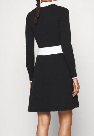 Kobieta ubrana w czarną sukienkę z długim rękawem, z białym paskiem w talii i białym kołnierzykiem, stoi tyłem, z warkoczem spływającym po plecach.