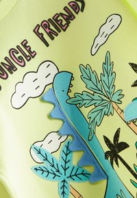 Lys grøn sweatshirt med et stort blåt dino-grafik, palmeblade, skyer og teksten "Jungle Friends" i fed pink. Blå teksturerede accenter.
