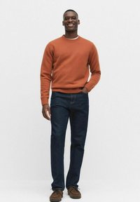 Rostfärgad stickad tröja med ribbade ärmar, blå jeans och bruna mockaskor. Modellen står mot en vit bakgrund och ler.