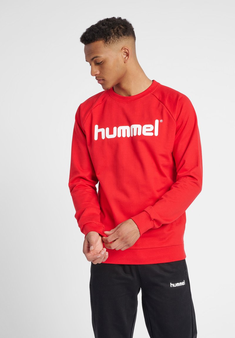Sudadera roja con cuello redondo y mangas largas, que presenta un gran logo blanco de "hummel" en la parte delantera. Combinada con pantalones negros.