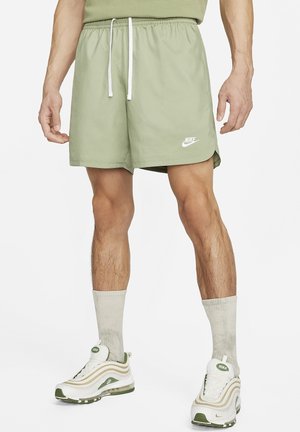 Homme portant un short Nike vert clair avec cordon blanc, des chaussettes beiges et des baskets Nike blanches et vertes, debout sur fond blanc.