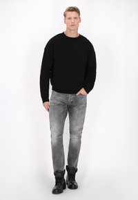 Sort bomulds-sweatshirt med rund halsudskæring, parret med slidte grå jeans. Modellen bærer sorte snørebåndsstøvler. Enkel, afslappet design.