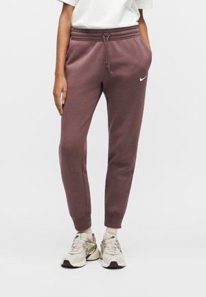 Osoba ubrana w purpurowe dresy Nike ze ściągaczem w pasie i białe sneakersy, stojąca z jedną ręką w kieszeni na jednolitym tle.