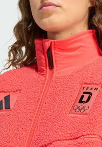 Coral fleece jack met hoge kraag, rits aan de voorkant en zwarte accenten. Heeft het Olympisch logo en een "TEAM D"-badge op de borst.
