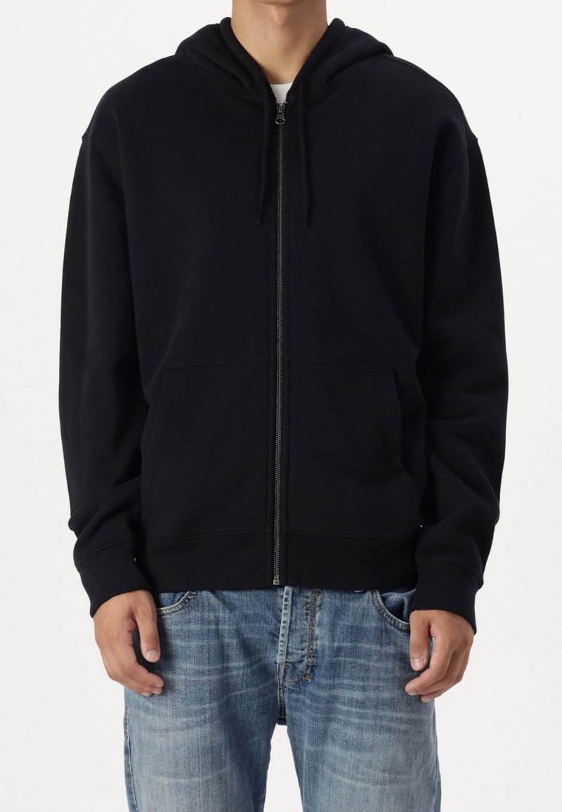 Sudadera con capucha negra con cremallera, bolsillos delanteros, capucha ajustable con cordón y puños acanalados. La tela parece suave con una textura lisa.