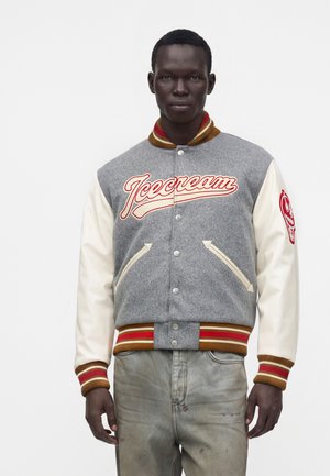 Mann trägt eine grau-weiße Collegejacke mit rot-gold gestreiften Bündchen und „Icecream“-Logo auf der Brust, kombiniert mit abgenutzten grauen Hosen.