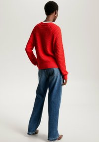 Personne portant un pull texturé rouge à manches longues et un jean ample bleu, se tenant avec une main sur la hanche, de dos.