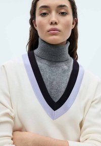 Vit tröja med bred V-ringning med svart och ljuslila kant, lager över en grå polotröja. Mjuk textur, avslappnad design.