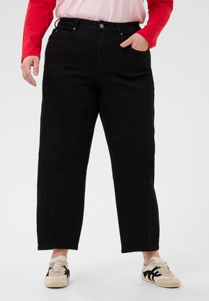 Personne portant un pantalon noir à jambes larges, une chemise rouge à manches longues et des baskets blanches avec des bandes noires, debout sur un fond uni.