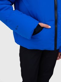 Osoba u plavoj jakni s rukom u džepu, crne hlače su vidljive, zatvarač jakne je djelomično prikazan, tekst "10k" na rukavu.