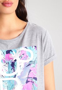 Camiseta gris de manga corta con un gráfico floral colorido en rosa y azul, que incluye un texto en blanco en el centro.