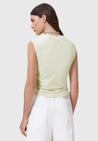 Top senza maniche di un verde chiaro con una texture liscia, dettagli arricciati sulle spalle e un sottile logo sul retro, abbinato a pantaloni bianchi.