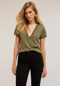 Top de punto de manga corta verde oliva con un escote en V profundo y ribetes en contraste, combinado con unos jeans ajustados negros.