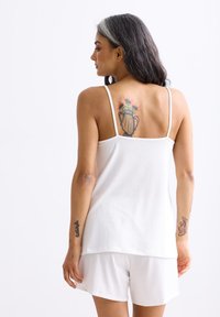 Witte mouwloze top met dunne banden, gecombineerd met witte shorts. Heeft een gladde textuur en een schoon ontwerp. Tattoos zichtbaar op de armen en rug.