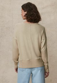 Maglione lavorato a maglia beige con ampio collo, spalle scese e fondo a costine. Caratterizzato da un dettaglio cucitura verticale e una texture morbida. Indossato con jeans azzurri chiari.