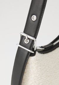 Curea din piele sintetică neagră cu accesorii din metal argintiu, atașată la o geantă din material texturat crem. Dispune de un închizător cu fermoar și detalii cu logo.