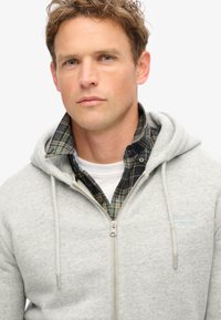 Grijze zip-up hoodie met trekkoorden, gedragen over een zwart-groene geruite shirt. Zachte textuur en subtiele branding op de borst.