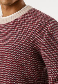 Roter und grauer gemusterter Strickpullover mit einem cremefarbenen gerippten Kragen. Der strukturierte Stoff zeigt ein diagonales Hahnentrittmuster. Nahaufnahme.