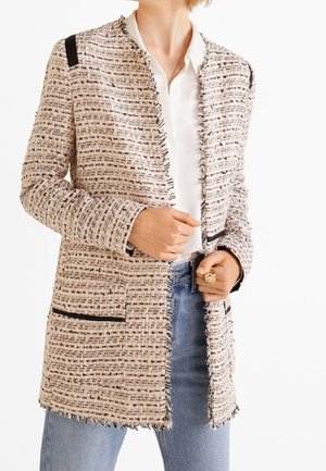 Femme portant une veste en tweed beige et noire avec des bords à franges par-dessus un chemisier blanc et un jean bleu clair, tenant le devant de la veste.