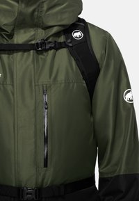 Grüne wasserdichte Jacke mit einem schwarzen unteren Abschnitt, ausgestattet mit Reißverschlüssen, verstellbaren Trägern und einem runden Logo am Ärmel.