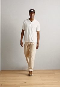 Cămașă polo albă, pantaloni bej, pantofi maro; persoană mergând pe podea din lemn, fundal minimalist. Șapcă cu logo.