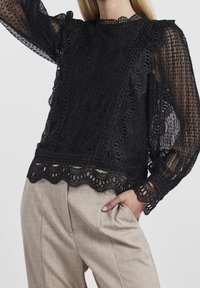 Sort bluse af blonder med intrikate mønstre og en bølget kant, parret med beige skræddersyede bukser, der viser en afslappet pasform og plisseringer.
