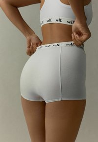 Valkoiset saumattomat shortsit, jotka on valmistettu pehmeästä kankaasta, joissa on leveä vyötärönauha, johon on painettu mustalla "self.". Suunnittelu on sileää ja tiukasti istuvaa.