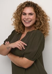 Olivovo zelená blúzka s nariasenými rukávmi a výstrihom do V. Modelka má kučeravé vlasy po plecia a tetovanie na vnútornej strane predlaktia.
